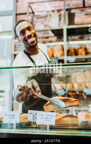 Lächelnd Mann bei der Arbeit in einer Bäckerei, die ein Stück des Kuchens mit einer Zange Stockfoto