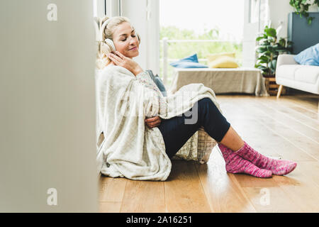 Frau eingehüllt in eine Decke am Fenster sitzen zu Hause hören von Musik mit Kopfhörer Stockfoto