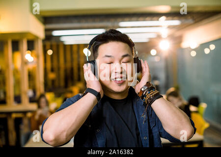 Jungen asiatischen Mann mit einem kabellosen Kopfhörer und höre die Musik, lächelnd und Stellung im modernen Studio Coworking Stockfoto