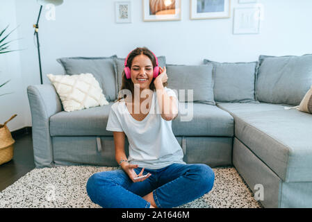 Glückliche junge Frau sitzt auf dem Boden im Wohnzimmer Musik hören mit Handy Stockfoto