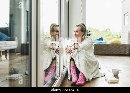 Frau eingehüllt in eine Decke am Fenster sitzen zu Hause Kaffee trinken Stockfoto