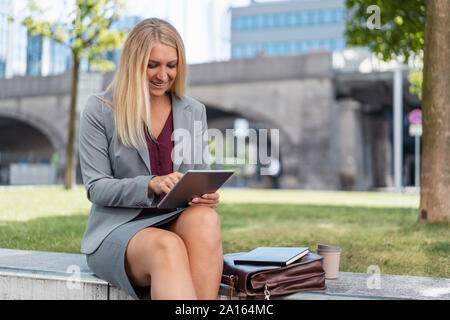 Lächelnde blonde Geschäftsfrau, sitzen auf einer Mauer im Freien mit digitalen Tablet Stockfoto