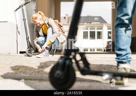 Junge Frau Schuhe binden, während Freund wartet mit e-Scooter Stockfoto