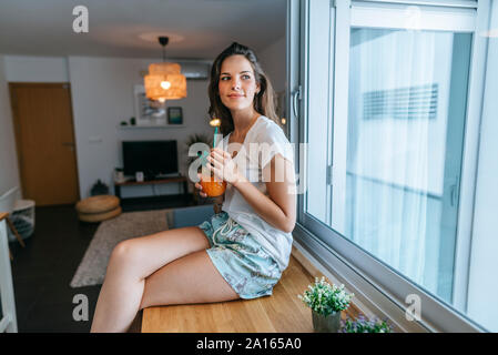 Lächelnde junge Frau sitzt auf der Arbeitsplatte in der Küche mit einem Getränk Stockfoto