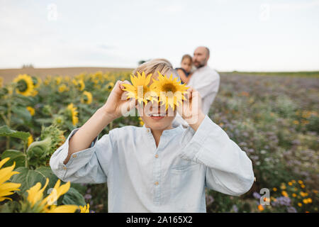 Verspielte Junge über seine Augen mit Sonnenblumen auf einem Feld mit einer Familie im Hintergrund Stockfoto