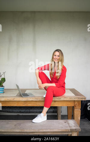 Lächelnde Frau mit Laptop sitzen auf hölzernen Tisch zu Hause Stockfoto