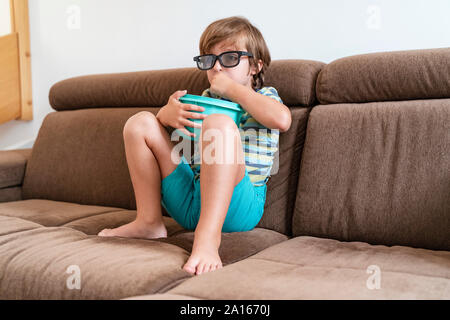 Junge sitzt auf der Couch zu Hause tragen 3D-Brille und essen Popcorn Stockfoto