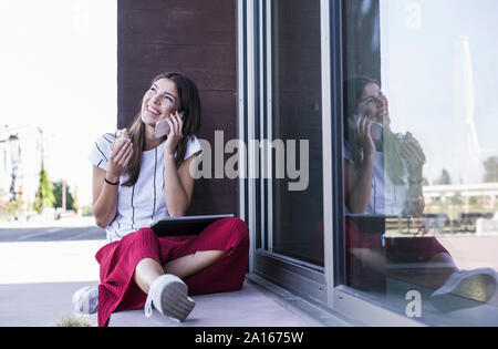 Junge Frau sitzt auf der Fensterbank an einem Gebäude mit Handy Stockfoto