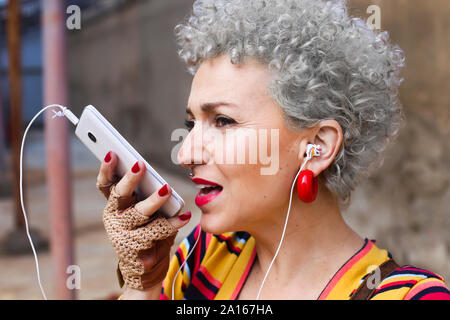 Portrait von durchbohrt, reife Frau mit grauen Locken mit Ohrhörer und Handy Stockfoto