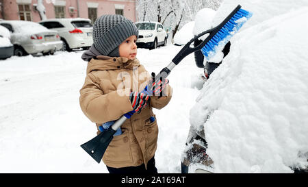 Closeup Bild des Jungen in beige Mantel reinigen das Auto nach dem Schneesturm mit Bürste Stockfoto