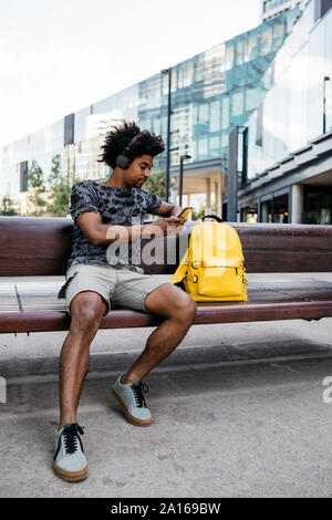 Mann auf der Bank sitzen Musik hören mit Kopfhörern und über Handy, Barcelona, Spanien Stockfoto