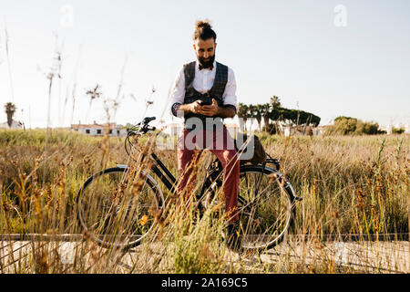 Gut gekleideter Mann mit seinem Fahrrad auf einem Holzsteg in der Landschaft mit Handy Stockfoto