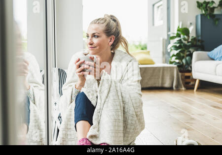 Frau eingehüllt in eine Decke am Fenster sitzen zu Hause Kaffee trinken Stockfoto