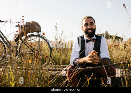 Gut gekleideter Mann sitzt auf einem Holzsteg in der Landschaft neben einem Fahrrad Stockfoto