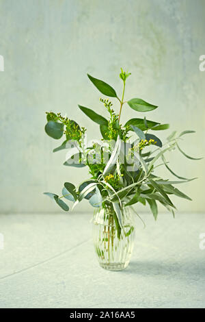 Blumenstrauß aus grünen Zweigen (Eukalyptus, wilde Oliven) und gelbe Blumen in glass vase Stockfoto