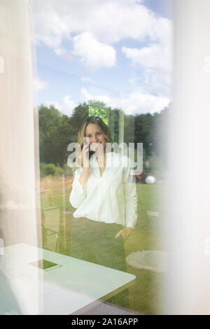 Lächeln, Geschäftsfrau, am Telefon zu sprechen zu Hause hinter Fensterglas Stockfoto