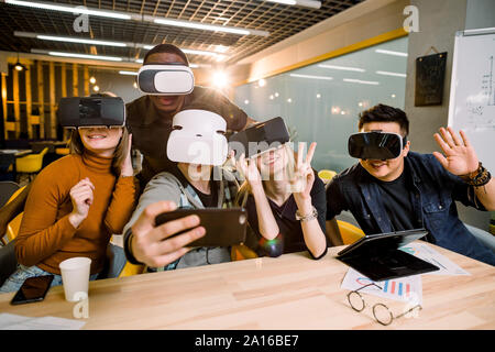 Fünf junge lustige Leute am Tisch vor sich sitzen, mit Virtual reality Brillen, Spaß und gestikulierend. VR-Brille Konzept Stockfoto