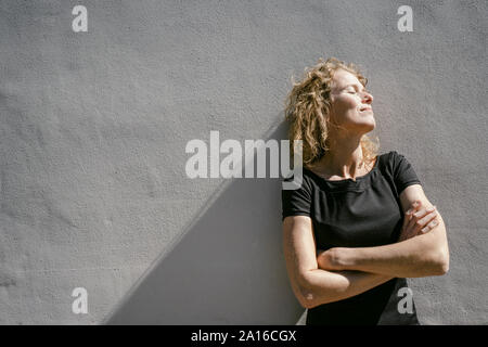 Lächelnd reife Frau mit geschlossenen Augen lehnte sich gegen graue Wand Sonnenlicht genießen. Stockfoto