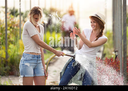 Zwei glückliche junge Frauen Spaß beim Blumen gießen mit Schlauch im Gewächshaus Stockfoto