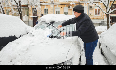 Der Mann in der Jacke versucht, dem Auto vom Schnee zu mit Bürste bei frostigen Morgen reinigen Stockfoto