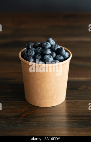 Frische organische Blaubeeren in einem Pappbecher. Dunkler Holztisch, hohe Auflösung Stockfoto