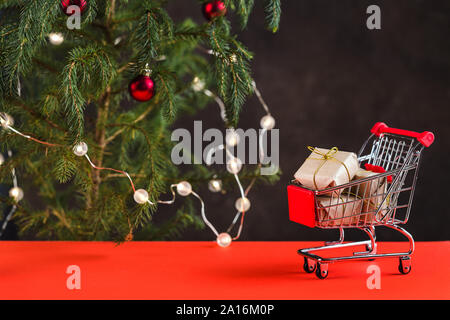 Frohes Neues Jahr 2020. Weihnachten Komposition. Mini Trolley mit geschenkboxen in der Nähe eines Weihnachtsbaumes mit Spielzeug auf dem roten Tisch Stockfoto