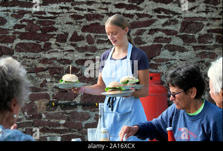 Junge Frau Kellnerin serviert Burger zu Kunden außerhalb Essen an den Tischen im Restaurant in Runwayskiln Marloes Pembrokeshire Wales UK KATHY DEWITT Stockfoto