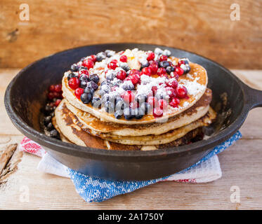 Stapel von hausgemachten oat Pfannkuchen mit Zucker, Blaubeeren und Preiselbeeren in Gusseisen Pfanne auf rustikalen Tisch. Soft Focus. Kopieren Sie Platz. Sprach Stockfoto