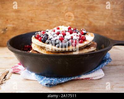 Stapel von hausgemachten oat Pfannkuchen mit Zucker, Blaubeeren und Preiselbeeren in Gusseisen Pfanne auf rustikalen Tisch. Soft Focus. Kopieren Sie Platz. Sprach Stockfoto