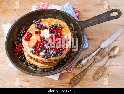 Stapel von hausgemachten oat Pfannkuchen mit Zucker, Blaubeeren und Preiselbeeren in Gusseisen Pfanne auf rustikalen Tisch mit alten Tafelsilber. Weich Stockfoto