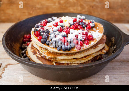 Stapel von hausgemachten oat Pfannkuchen mit Zucker, Blaubeeren und Preiselbeeren in Gusseisen Pfanne auf rustikalen Tisch. Soft Focus. Kopieren Sie Platz. Sprach Stockfoto