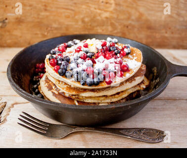 Stapel von hausgemachten oat Pfannkuchen mit Zucker, Blaubeeren und Preiselbeeren in Gusseisen Pfanne auf rustikalen Tisch mit alten Tafelsilber. Weich Stockfoto