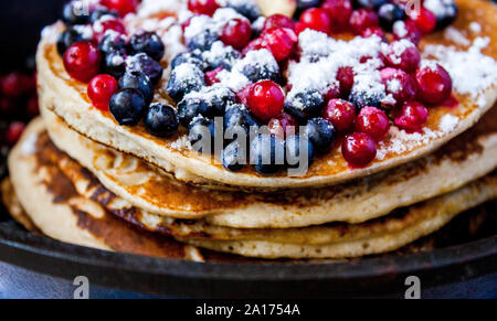 Stapel von hausgemachten oat Pfannkuchen mit Zucker, Blaubeeren und Preiselbeeren in Gusseisen Pfanne auf rustikalen Tisch. Soft Focus. Kopieren Sie Platz. Sprach Stockfoto