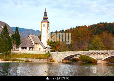 Bohinj See und der Kirche von Johannes dem Täufer und der Glockenturm und der Julischen Alpen mit Wald im Herbst Farben. Nationalpark Triglav, Slowenien. Stockfoto