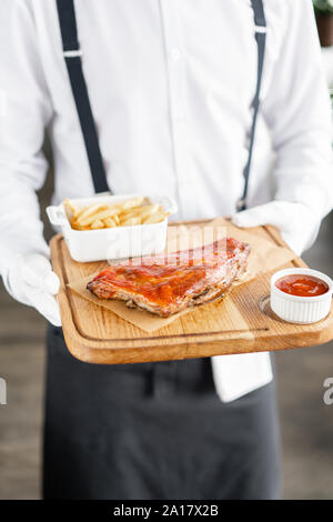 Der Kellner ist eine hölzerne Platte leckere Rippchen. Full Rack der RIBS BBQ auf Holzplatte mit Pommes Frites und Salat Stockfoto