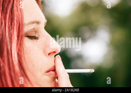 Closeup portrait einer hübschen frau mit geschlossenen Augen, Ausspucken von Zigarettenrauch in einem Park mit viel Grün im Hintergrund Stockfoto