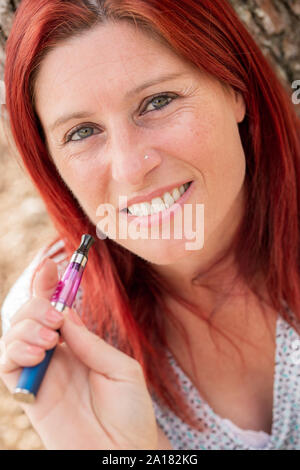 Portrait einer hübschen rothaarigen Frau gegen einen Baum Rauchen eine elektronische Zigarette in einem Park schiefen Stockfoto