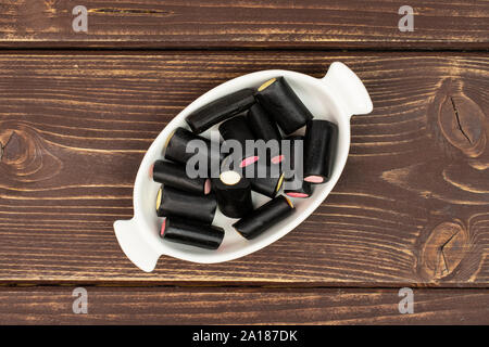 Menge ganz süßen schwarzen Lakritz Bonbons in weiße, ovale Keramik Schüssel flatlay auf braunem Holz Stockfoto