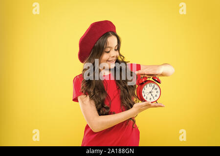 Glückliches Mädchen mit langen Locken in Baskenmütze. Kind mit Wecker. Zeitlose Mode. Pariser Kind auf Gelb. Schönheit Friseur. Kleines Mädchen im französischen Stil hat. Mädchen zeigt die Zeit auf der Uhr. Stockfoto