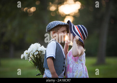 Kleine Mädchen küssen kleine Junge auf die Wange, während er hinter seinem Rücken versteckt Blumenstrauß Stockfoto