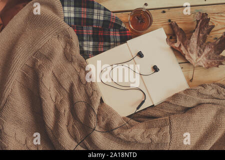 Herbst Hintergrund. Buch, Blatt und schwarz Ohrhörer auf einen gestrickten Pullover geöffnet. Die gestrickten Schal auf einen hölzernen Tisch. Herbst Wochenende Konzept. Ansicht von oben Stockfoto