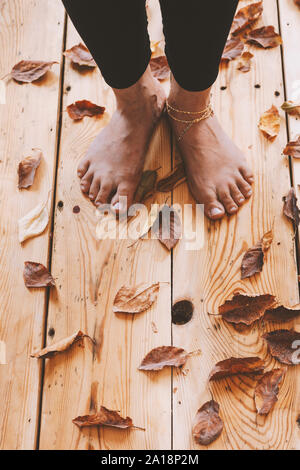 Die Füße der Person auf dem Holzboden mit Herbstlaub bedeckt. Herbst Konzept mit kopieren. Holzhaus, entspannen im warmen Herbst Stockfoto