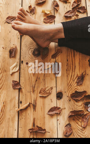 Die Füße der Person auf dem Holzboden mit Herbstlaub bedeckt. Herbst Konzept mit kopieren. Holzhaus, entspannen im warmen Herbst Stockfoto