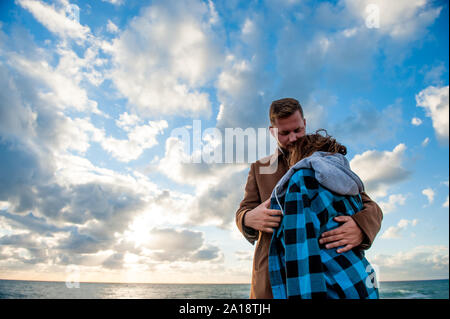 Liebe und Fürsorge Konzept junges Paar Mann im Mantel seiner zitternden Frau in Plaid Shirt und Hoodie in der Nähe von blauen Himmel mit Wolken und Meer umarmt Stockfoto