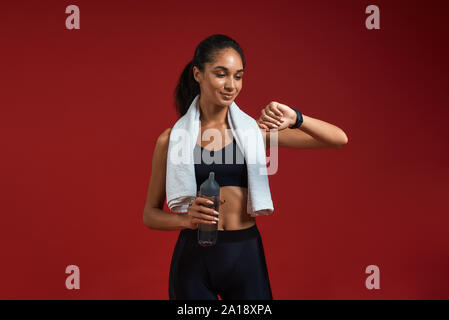 Wie spät ist es Junge afro amerikanische Frau in Sportkleidung mit Handtuch auf den Schultern hält Flasche Wasser und die Zeit zu überprüfen, während sie vor rotem Hintergrund stehen. Sportkonzept. Aktiver Lebensstil Stockfoto