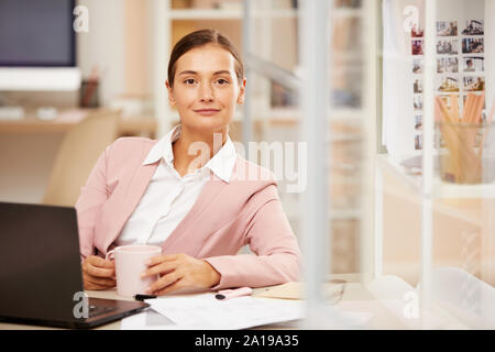 Portrait von selbstbewussten jungen Geschäftsfrau, Lächeln auf die Kamera beim Sitzen am Schreibtisch und Kaffee trinken Stockfoto