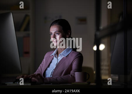 Junge zuversichtlich Geschäftsfrau am Tisch vor computer monitor sitzen und Tippen auf der Computertastatur Sie bis spät in die Nacht arbeiten Stockfoto