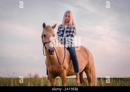 Junge blonde Mädchen in Blue Jeans und ein kariertes Hemd sitzt auf einem Pferd auf der Ranch closeup gekleidet Stockfoto