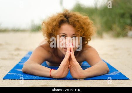 Junge rothaarige Frau mit Händen in Namaste liegt auf Yoga Matte am Strand Stockfoto