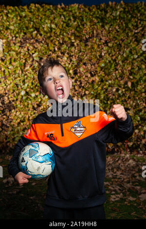 Ein acht Jahre alter Junge trägt eine Fußball-Streifen und halten ein Fußball unter dem Arm, mit der anderen Hand in eine Faust. Stockfoto